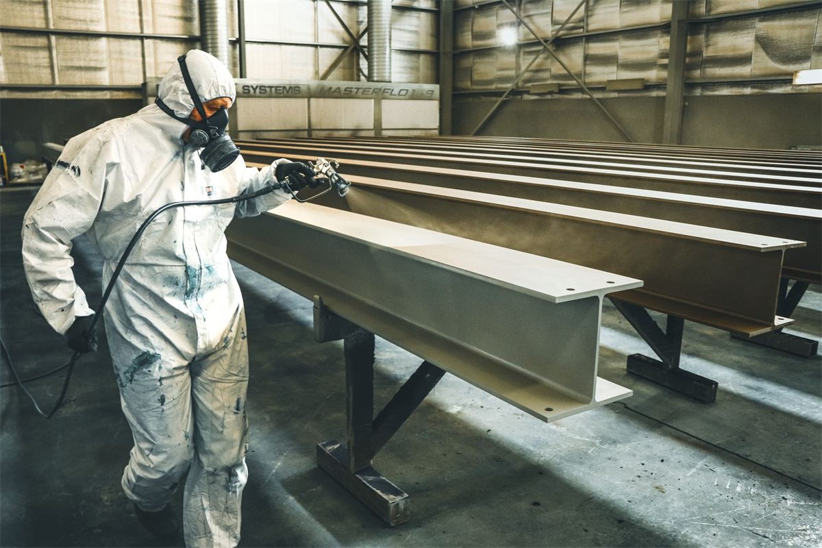 Painter in a white hooded coverall, half-face respirator and gloves spraying steel beams in a coating shop