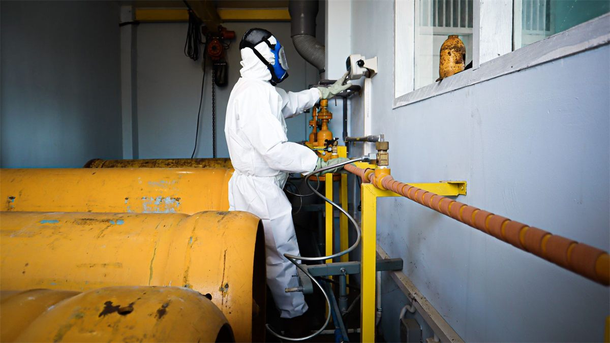 Worker in a white hooded coverall, full-face respirator and gloves operating valves beside yellow gas cylinders