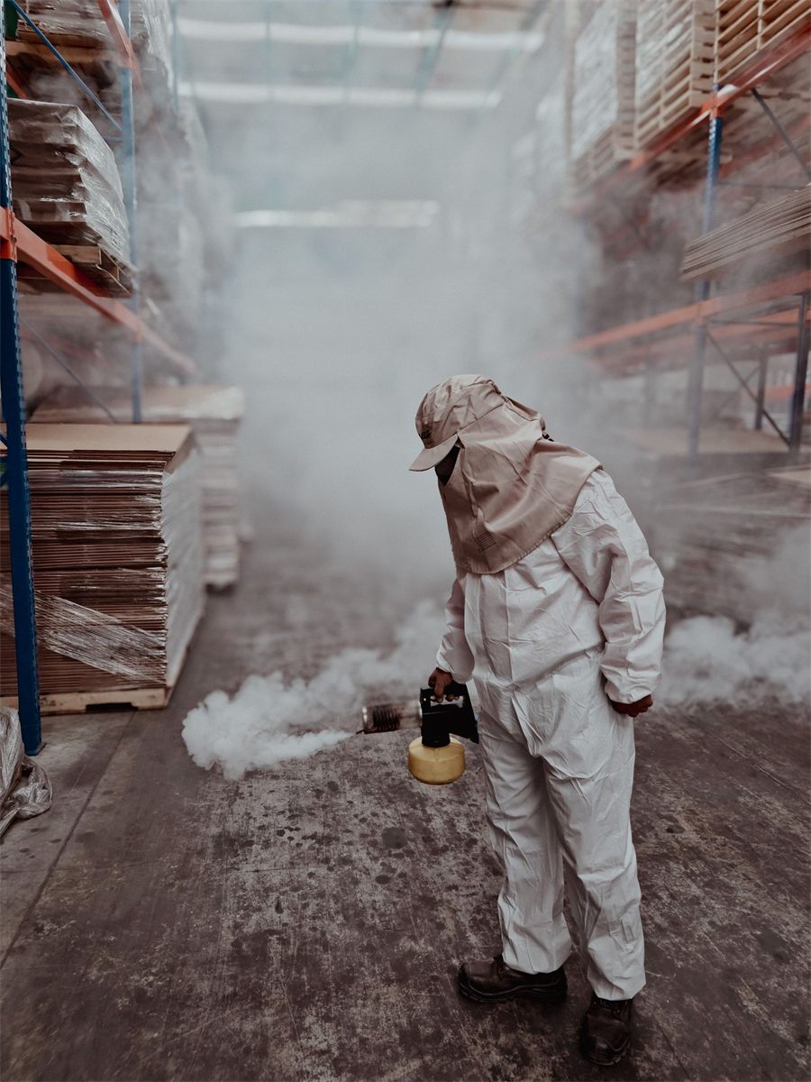 Pest control technician in a white coverall, cap with neck flap and boots fogging a warehouse aisle